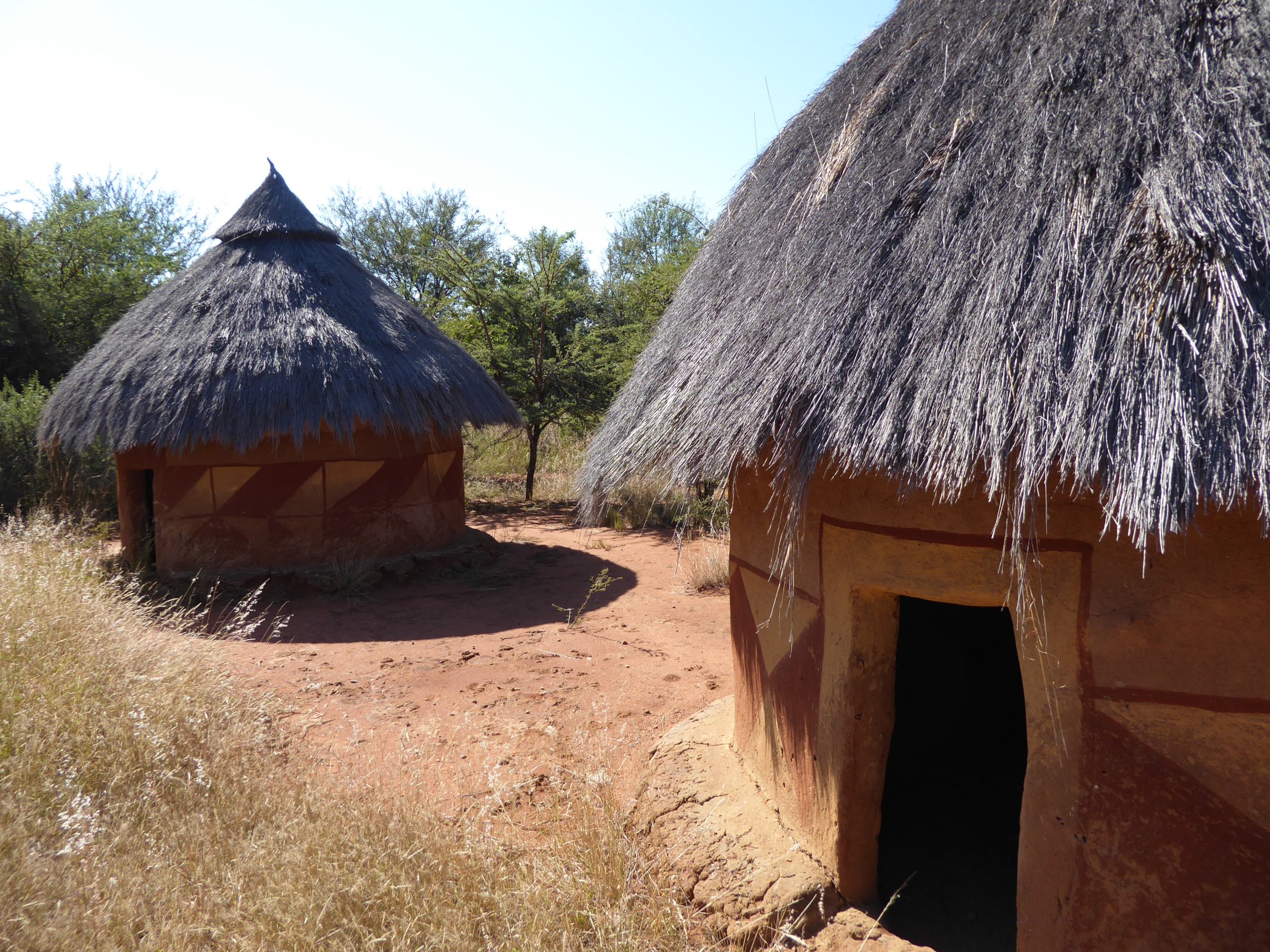 Two huts with straw roofs. The huts are cylindrical with a pointy roof. The huts are in a desert area with trees in the background.