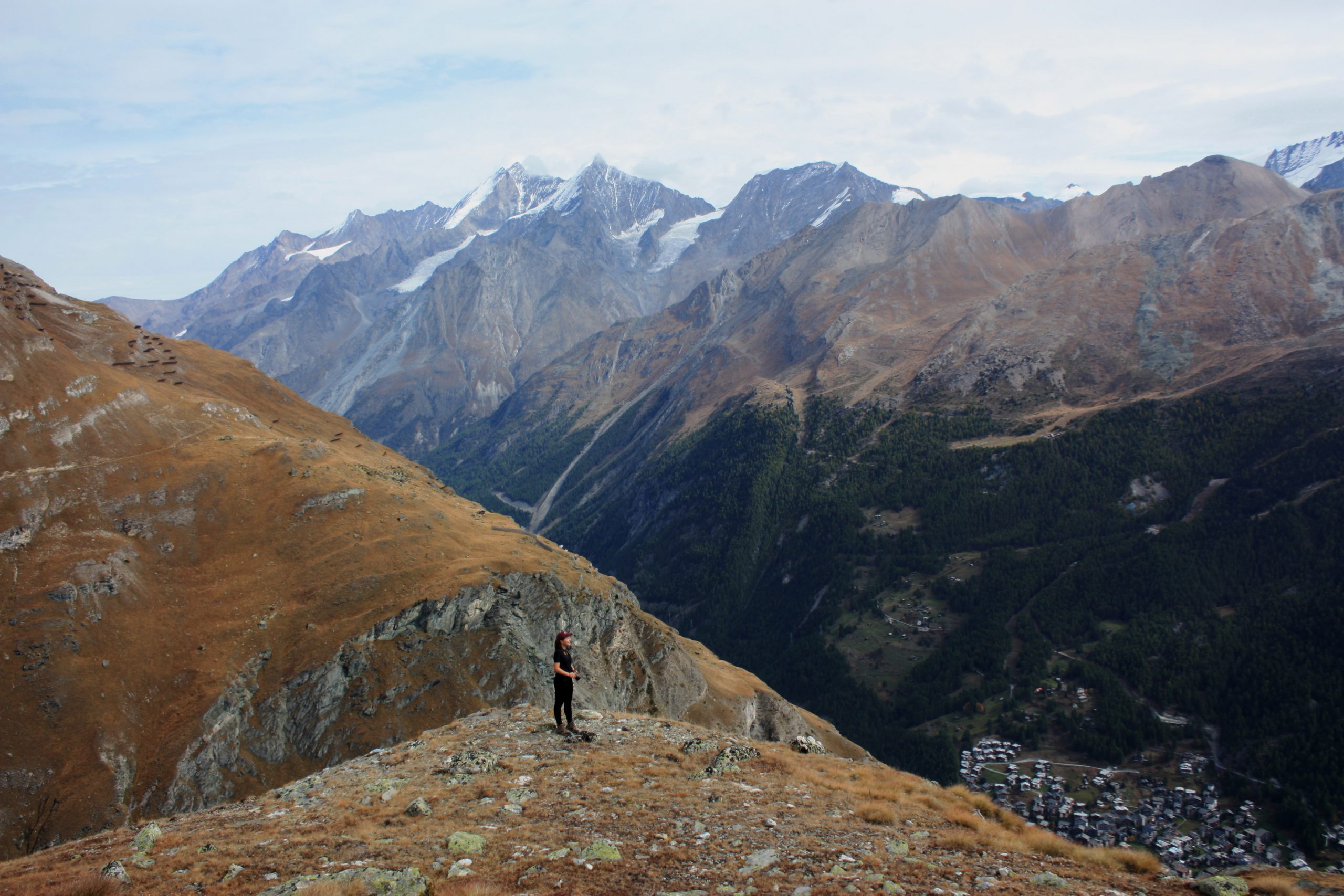 Student on a mountain with taller mountains in the background. The mountains are covered in trees, snows, and rocks. A small village can be seen in the bottom right of the photo, in a nearby valley.
