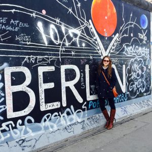Student abroad, standing in front of a mural with graffiti that reads: Berlin. She is wearing a blue coat and black pants with brown boots, bag, and glasses.