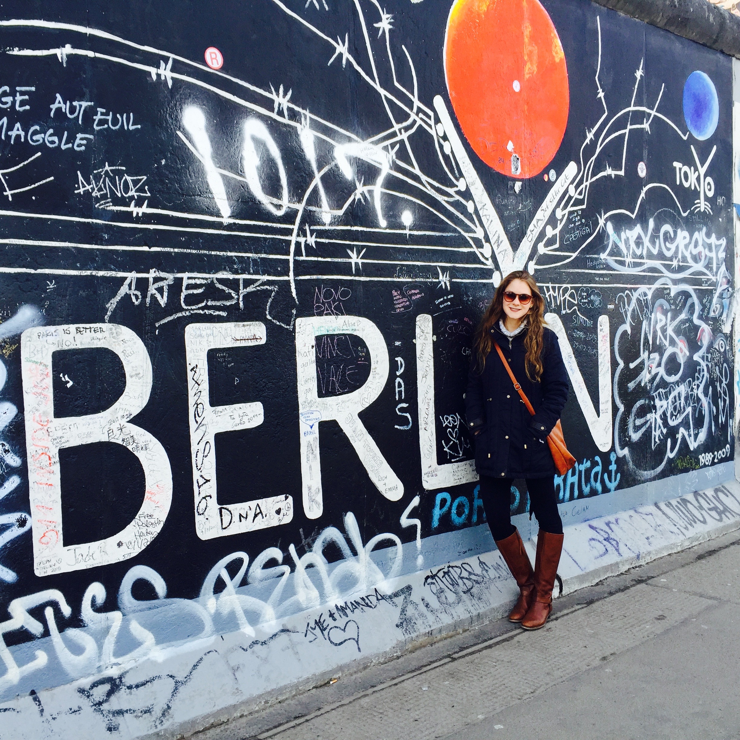 Student abroad, standing in front of a mural with graffiti that reads: Berlin. She is wearing a blue coat and black pants with brown boots, bag, and glasses.