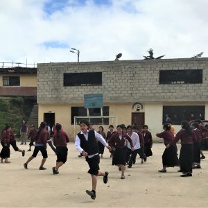 Children playing outside at school. They are wearing maroon, white, and black uniforms, the school building is behind them, and they are running around for recess.