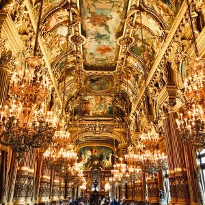 Inside of a mansion in Italy. The gilded decor includes ornate gold trims, paintings on the ceilings, large chandeliers, and tourists walking the palace.