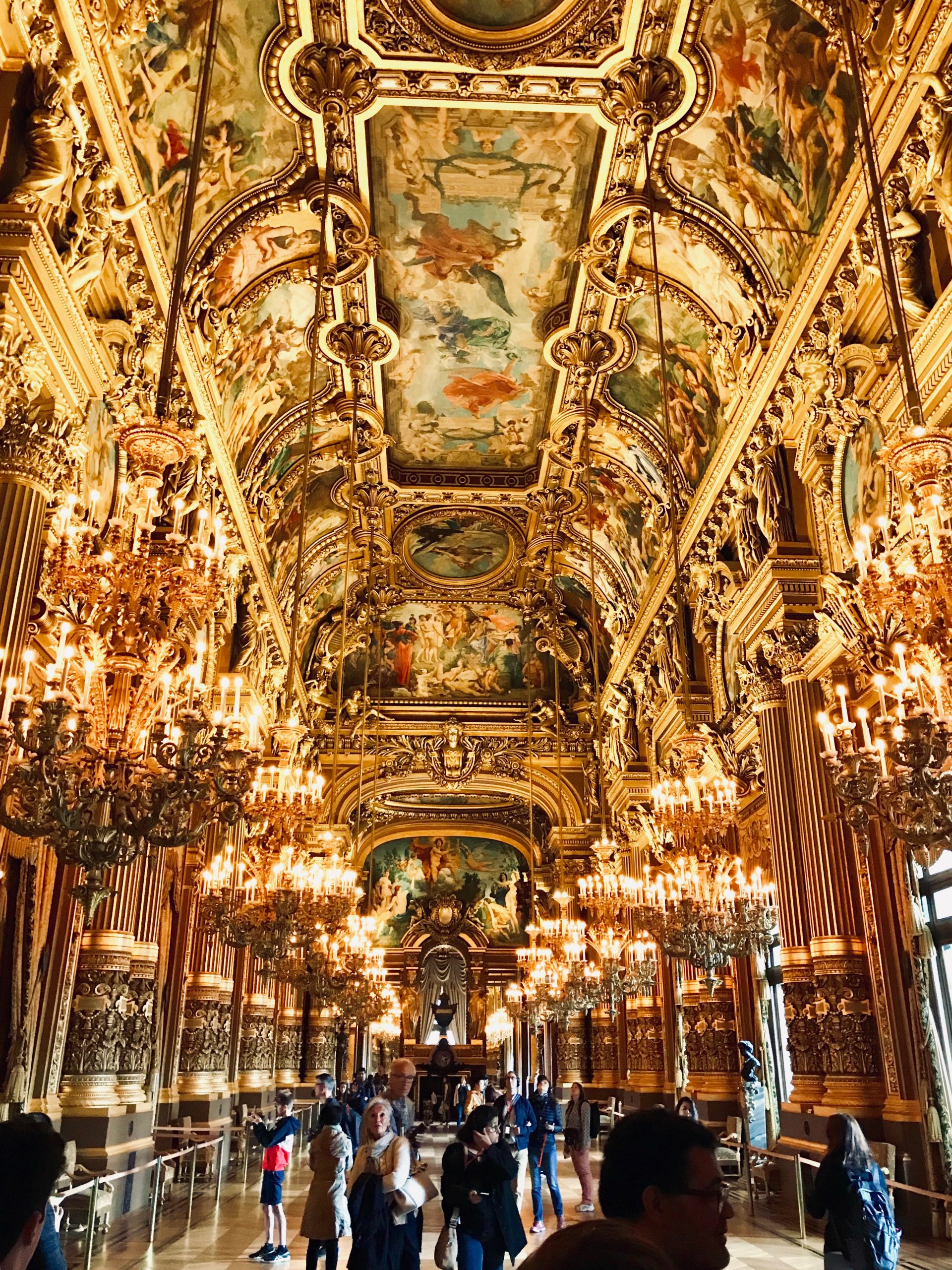 Inside of a mansion in Italy. The gilded decor includes ornate gold trims, paintings on the ceilings, large chandeliers, and tourists walking the palace.