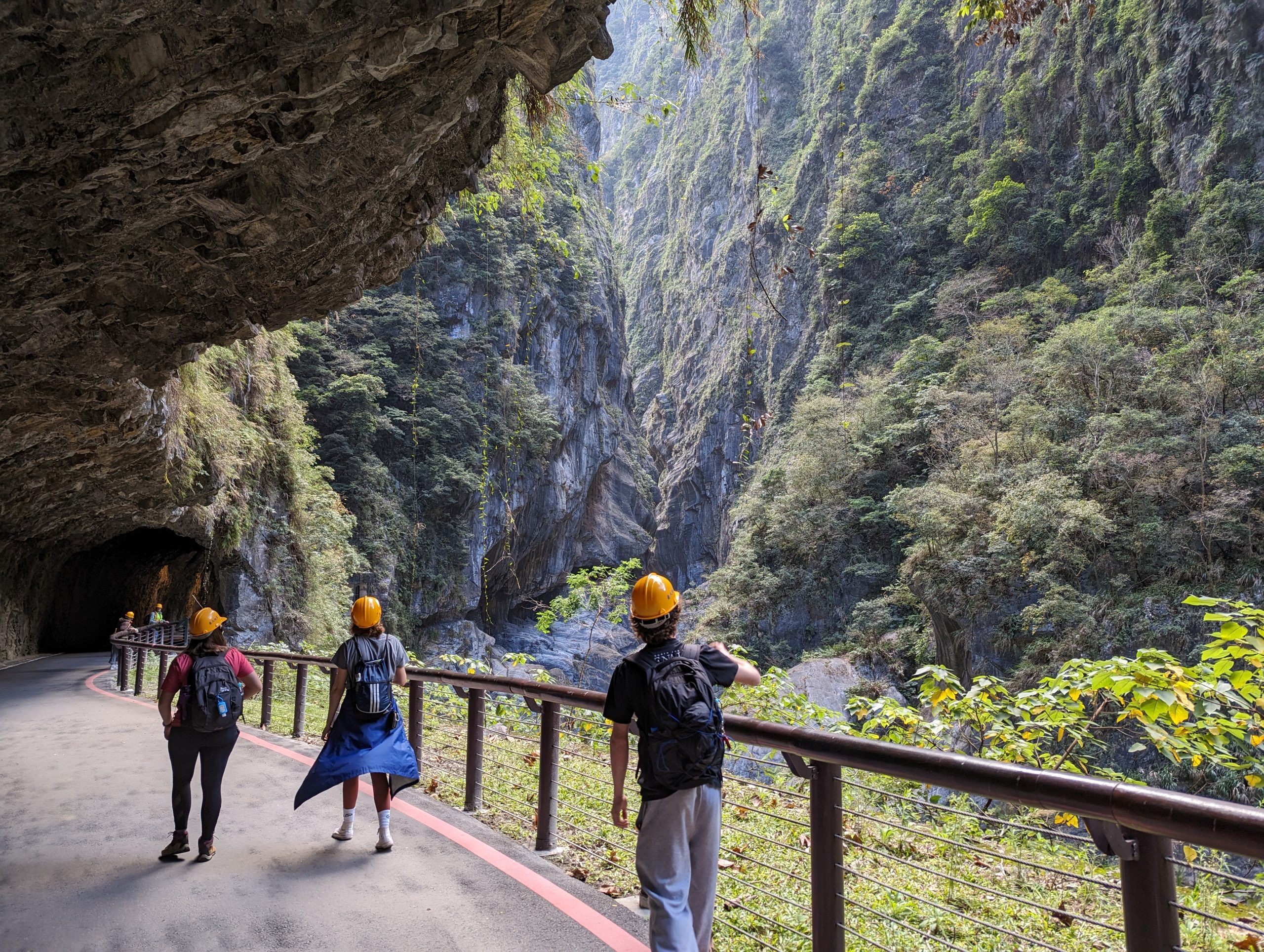 Students walking along a path with a railing, in yellow hard hats. The students are posed looking up at a mountain, and the path they are following leads into a cave within the mountain.
