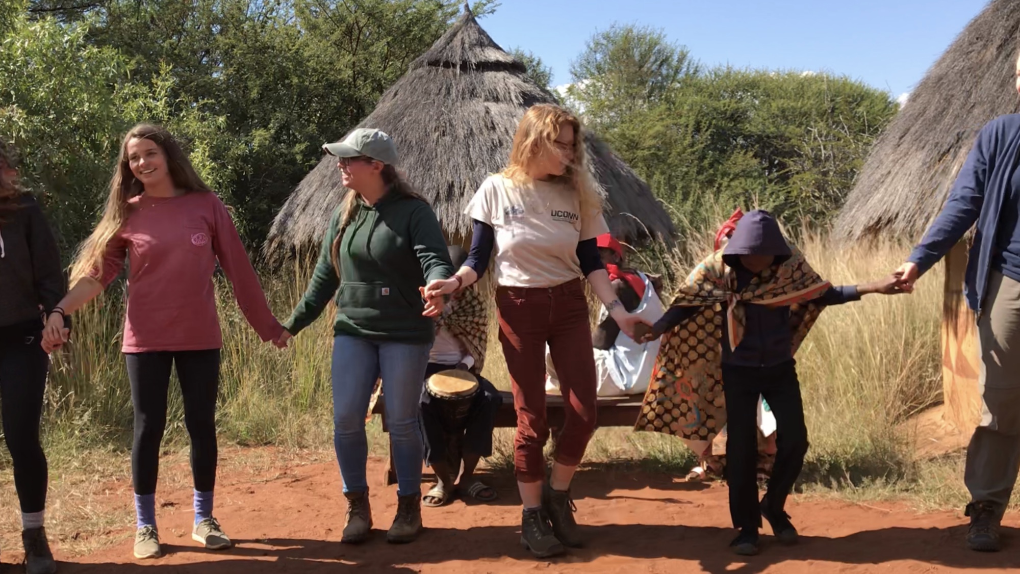 UConn students holding hands in front of a hut in a desert area with trees in the background abroad.