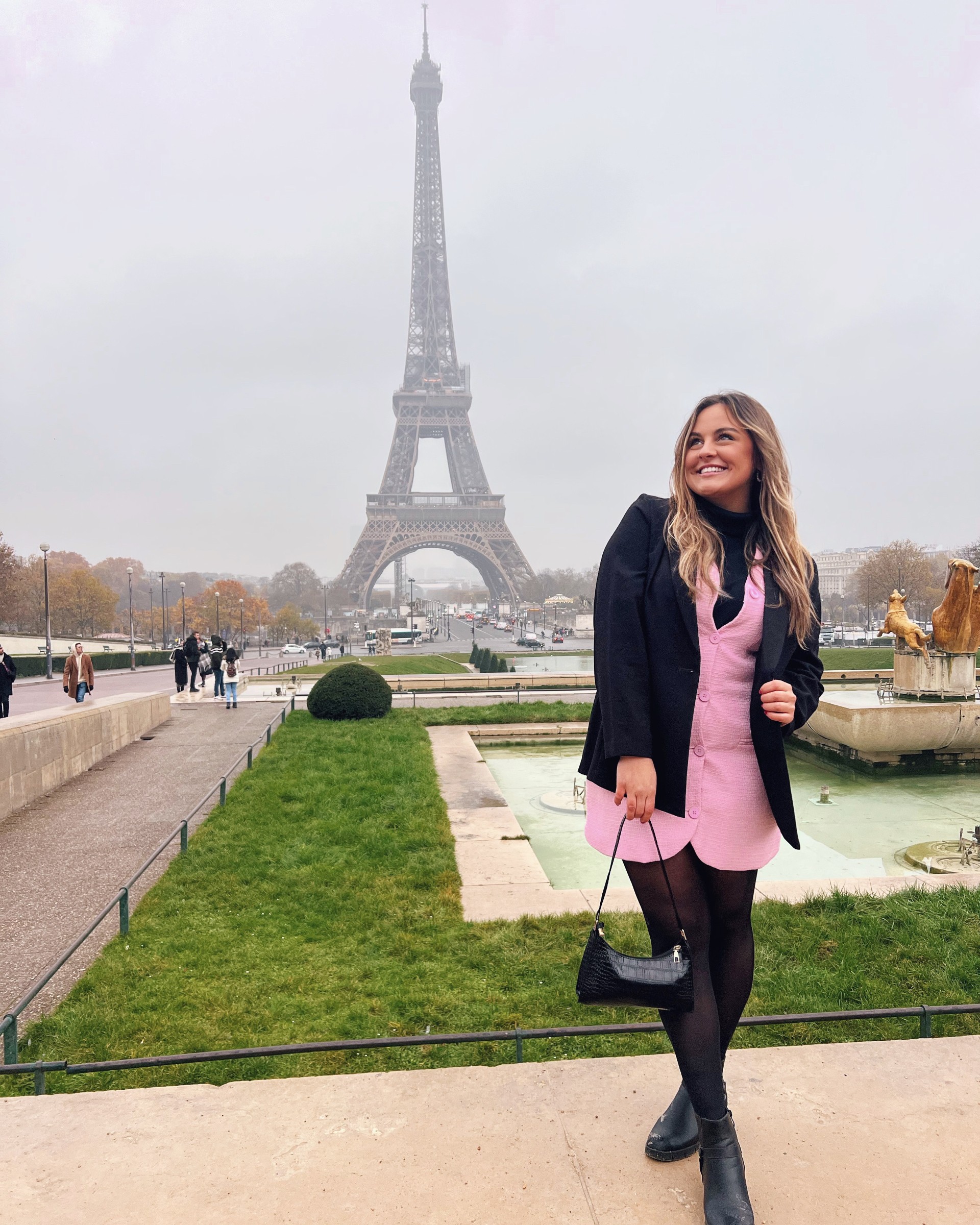 Student standing in front of the Eiffel tower on a gloomy day. She has blonde hair, is wearing a pink dress and blazer, and is holding a black bag.