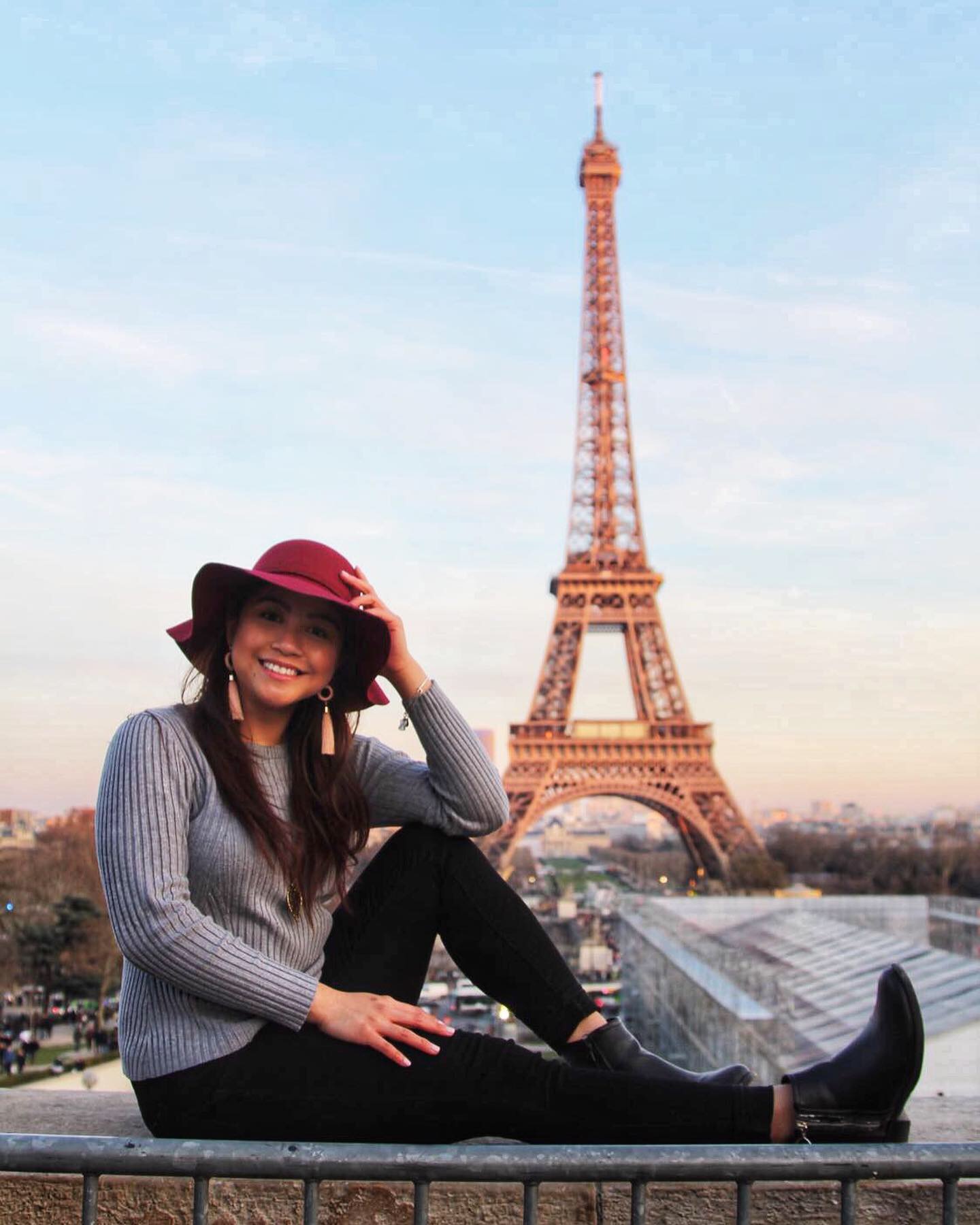 Student in a grey sweater, red hat, black pants and boots, and tan tassel earrings. She is sitting posted with her hand on her head, and he leg out in front of the Eiffel tower.