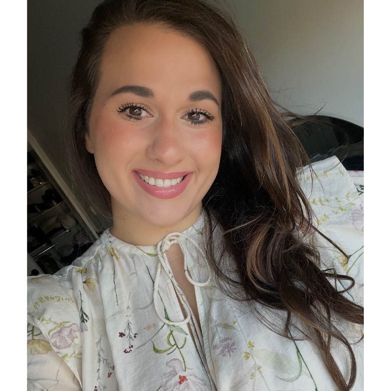 Photo of a white woman with long brown hair wearing a floral white blouse smiling at the camera.