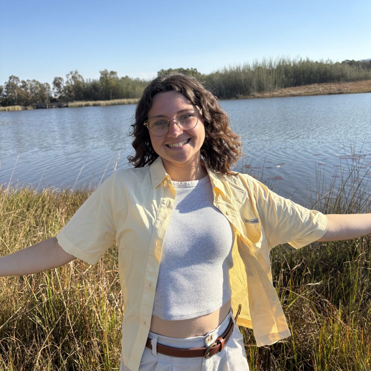 Female student wearing a yellow button up shirt with a white top underneath smiling with her hands out in front of a pond.