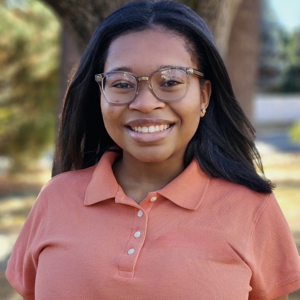 Photo of a smiling young black woman with medium length straight black hair wearing glasses and a pink collared short sleeve shirt.