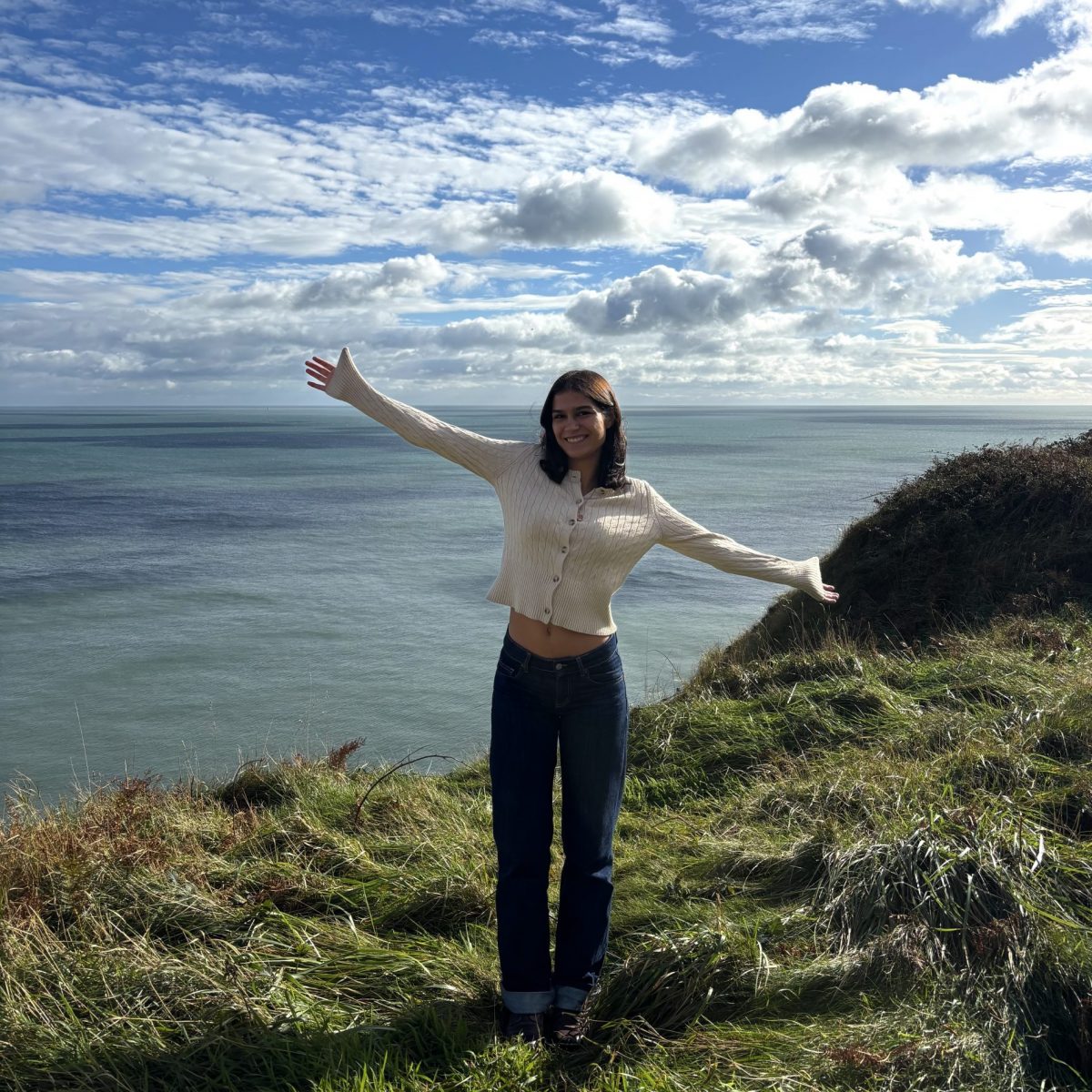 Female student wearing a white cardigan and jeans with her hands out on a grassy patch in front of the sea.