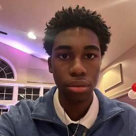 Photo of a young black man wearing a blue quarter zip with a silver cross and collared shirt underneath smiling at camera.