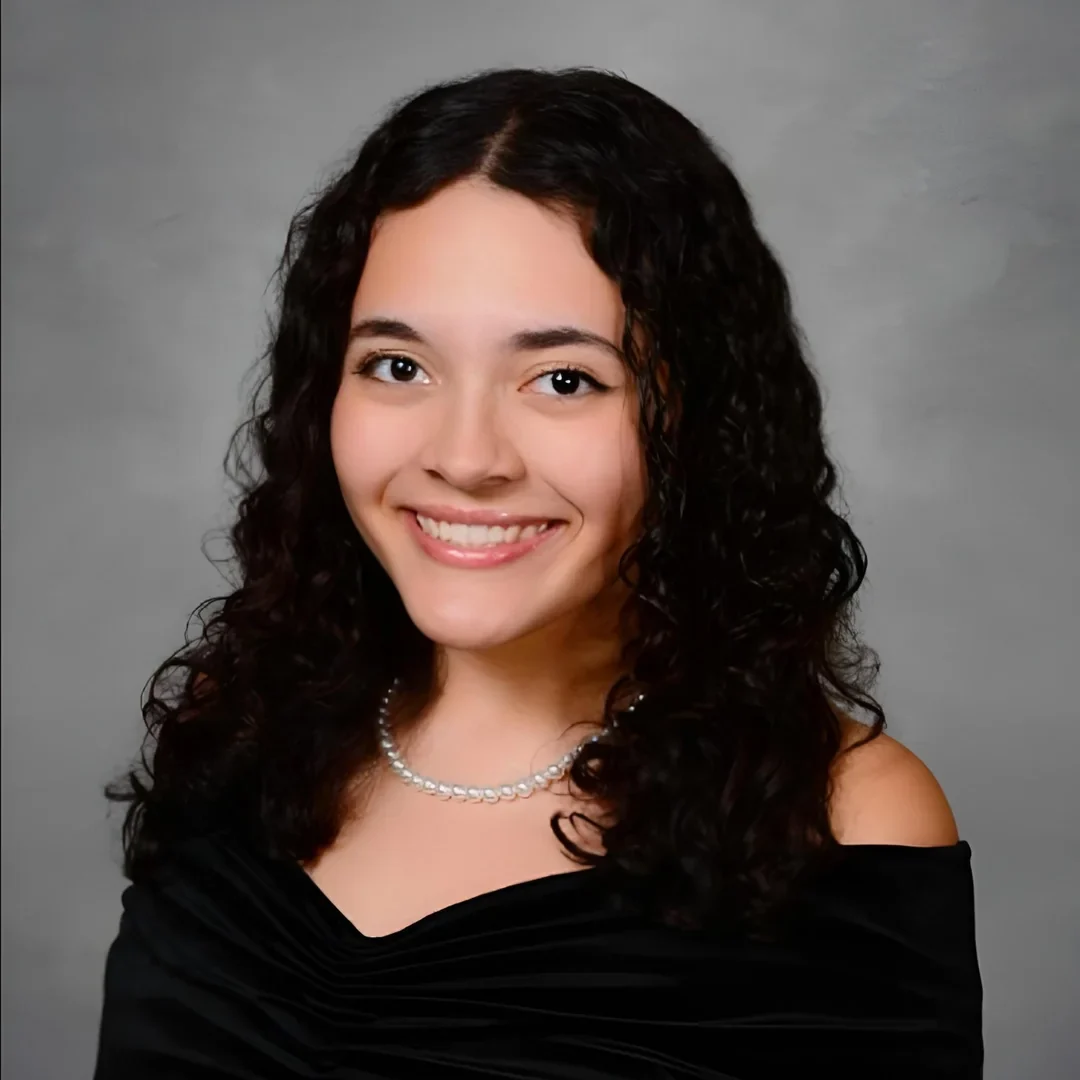 UConn student smiling at camera. She is a wearing a black V-neck off the shoulder top and has medium length black curly hair.