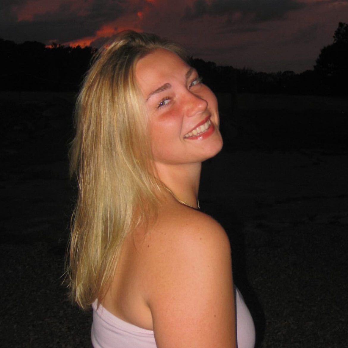 UConn Student smiling at camera wearing a pink tube top. She has medium length blonde hair and is posing in front of a red sunset.