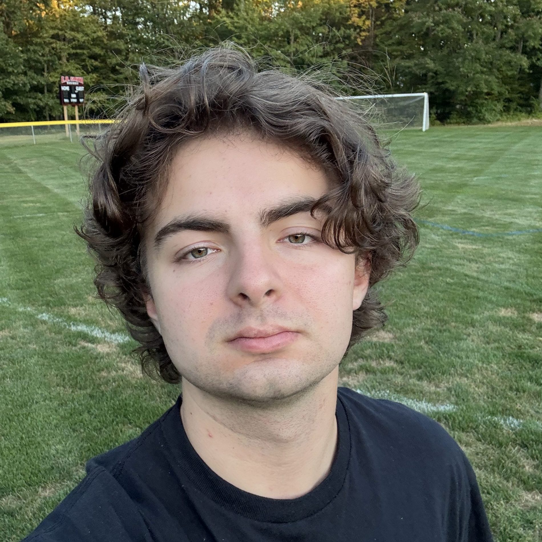 Male student with curly brown hair poses for selfie in a soccer field, wearing a black t-shirt.