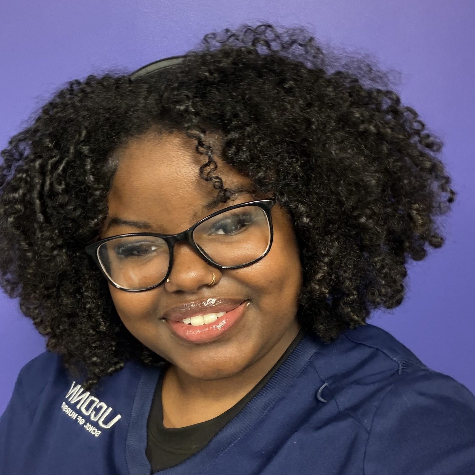 UConn student wearing nursing scrubs shift, black rimmed glasses, with short curly black hair smiling for camera.