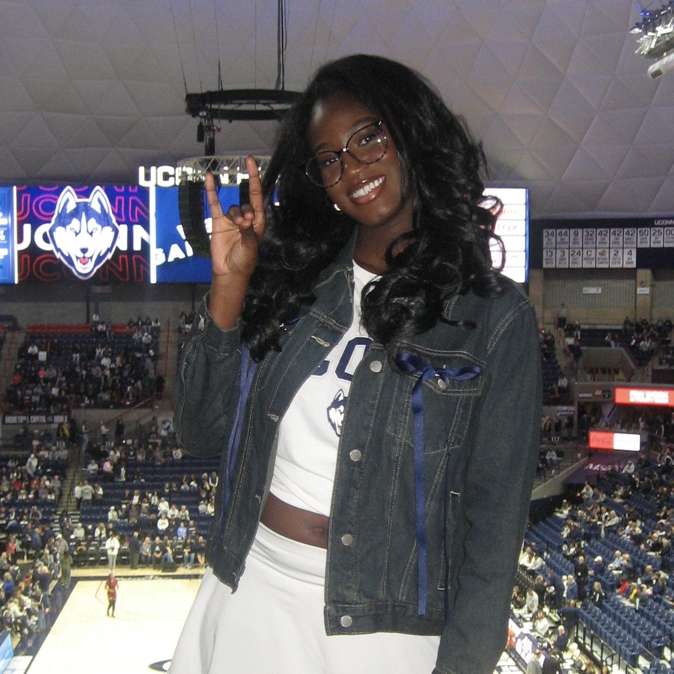 Female student with curled black hair smiles at a UConn Basketball game, holding up a husky hand sign while wearing a white skirt, white UConn shirt, and a customized denim jacket.
