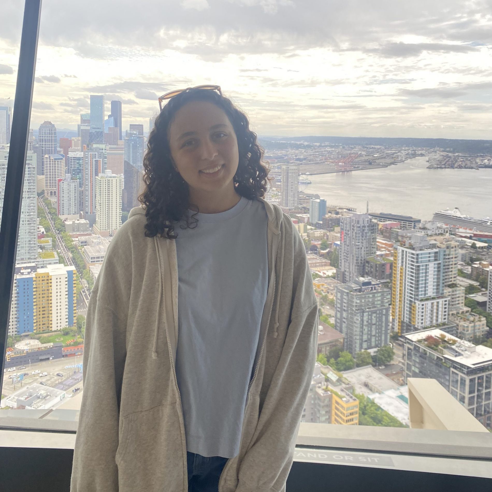 Student with shoulder legnth curly hair smiles standing in front of a window with a cityscape behind her, while wearing a plain t-shirt with a sweatshirt over it.
