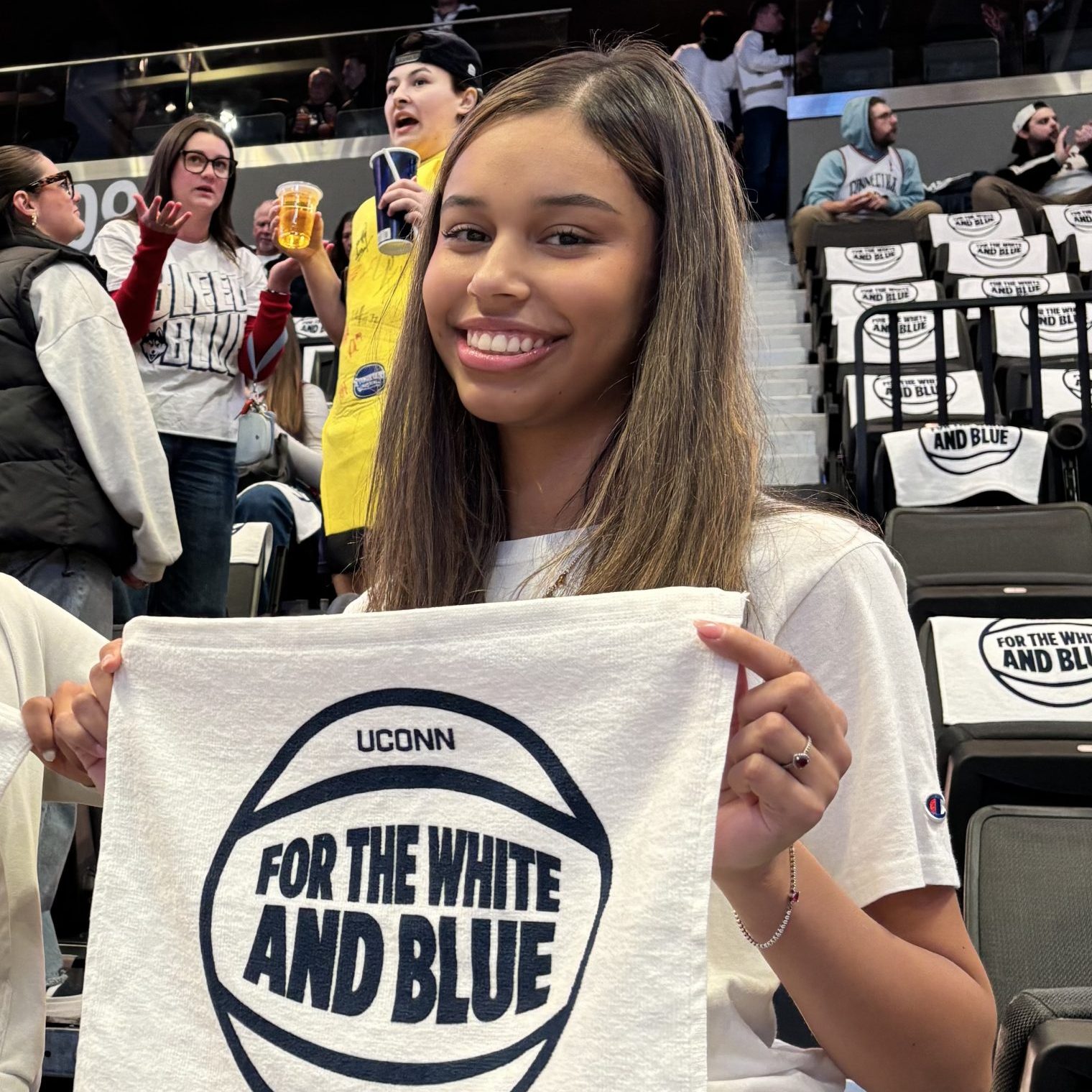 Female student with brown hair smiles while holding up a white UConn towel with the text: "For the White and Blue" inside a basketball graphic.