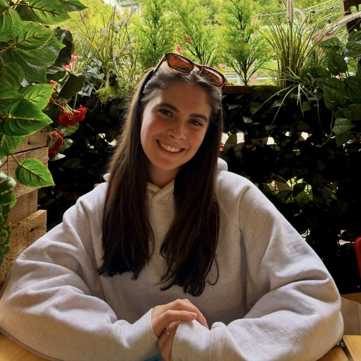 Female student with long brown hair pushed back by brown sunglasses and wearing a gray sweatshirt smiles with her hands in front of her surrounded by greenery.