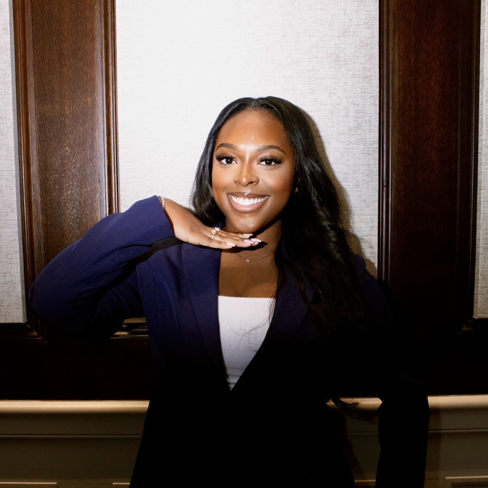Female student with curled black long hair smiles, holding one hand up under her chin while wearing a navy pantsuit.