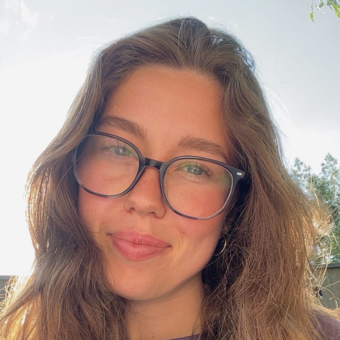 Female student with long brown hair smiles for camera while wearing black rimmed glasses