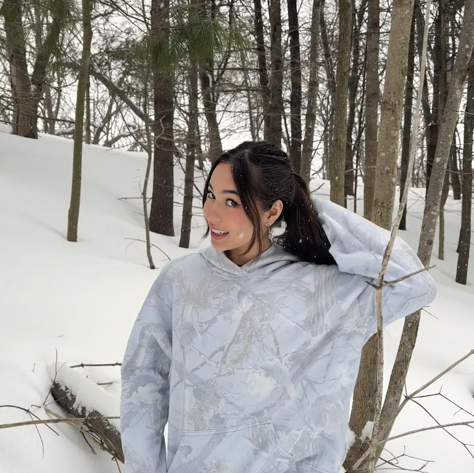 Female student poses for photo wearing a light gray camo style hoodie with her hair tied back in a snowy forest.