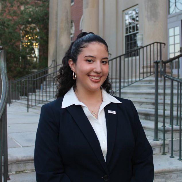 Female student with long curly black hair slicked back smiles wearing a black blazer and white collared shirt underneath.