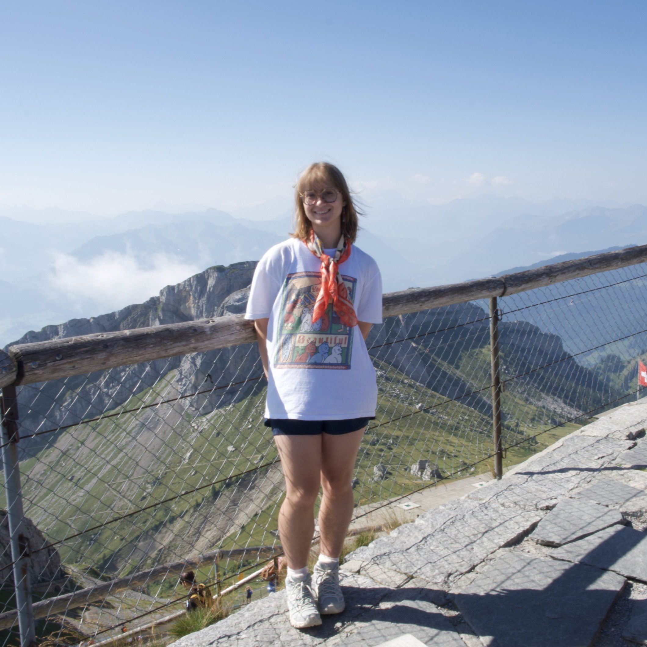 Female student with shoulder length hair wearing a t-shirt and shorts, standing in front of a railing with a valley and mountains visible behind her.