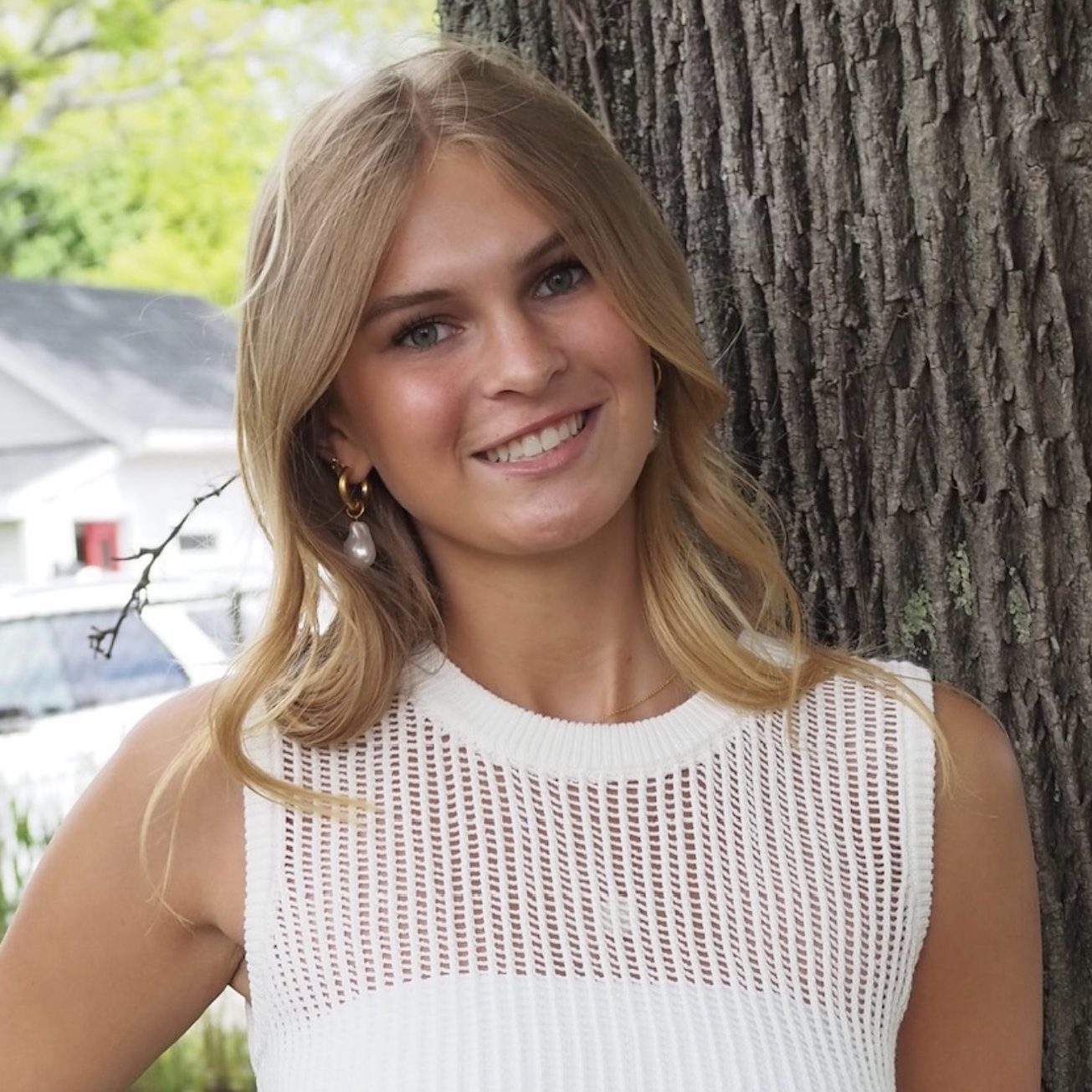 Female student with shoulder legnth blonde hair smiles for camera while leaning against a tree wearing a white sleeveless sweater.
