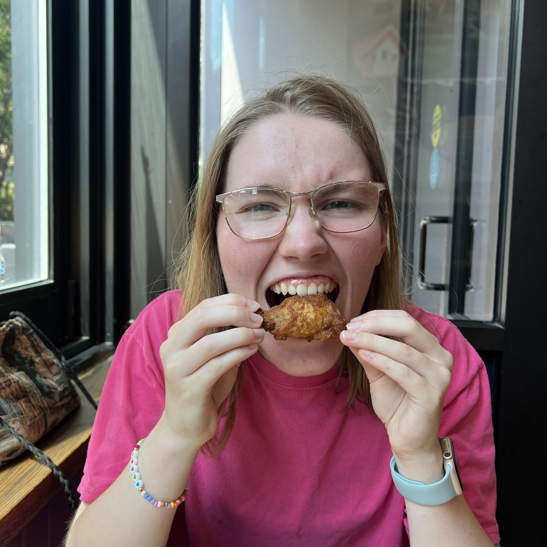 Student biting down on chicken wing in a restaurant setting while wearing a pink t-shirt.