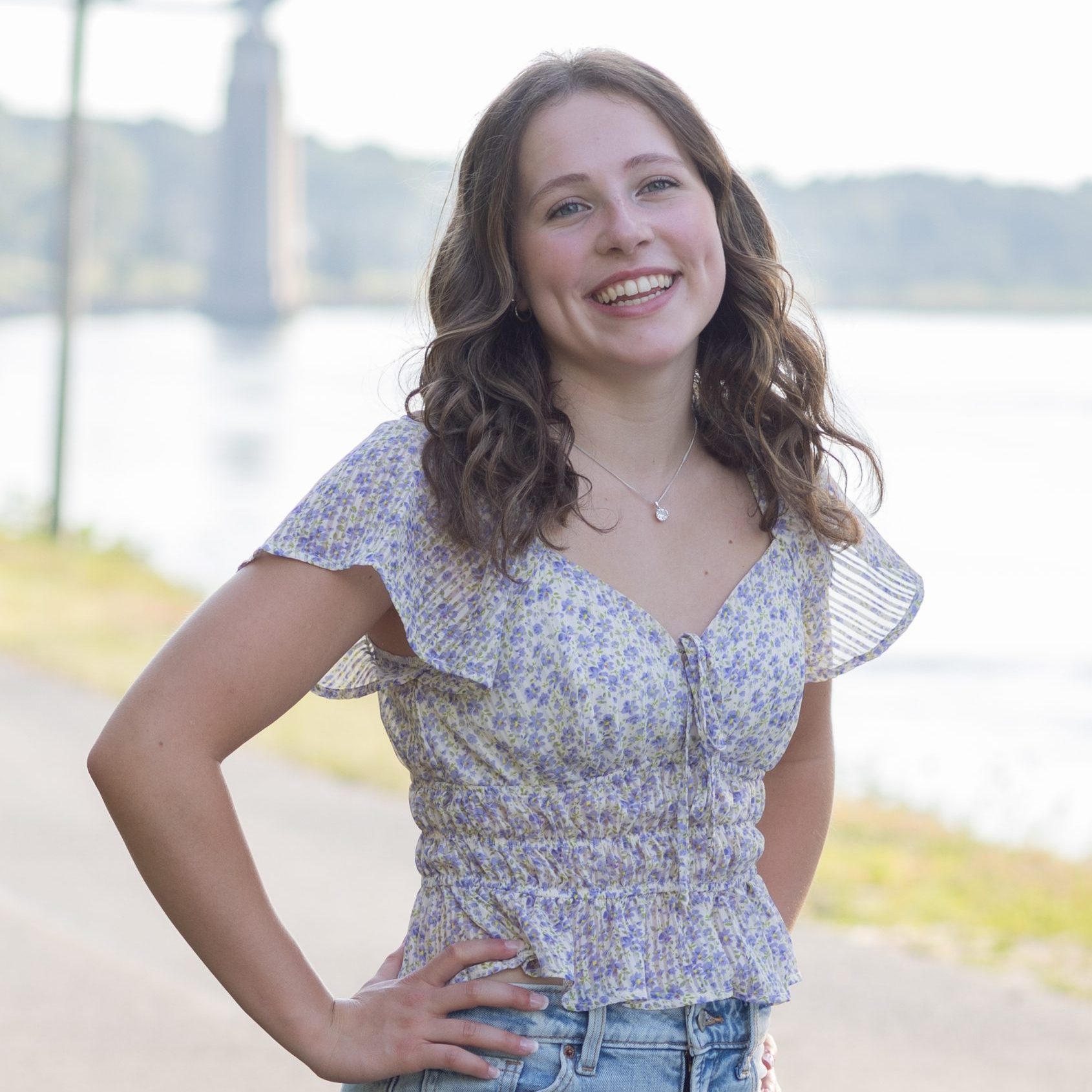Female student photo wearing a blue floral blouse and jeans, standing in front of a lake.