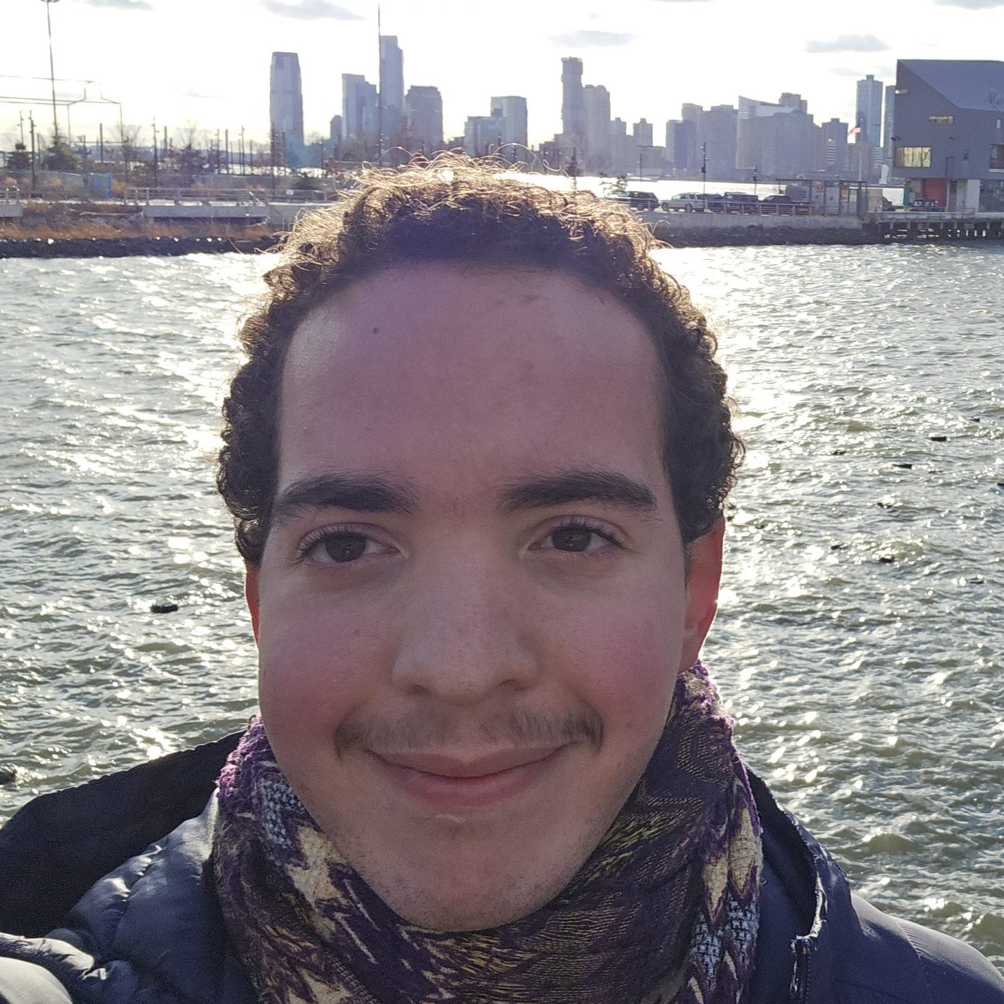 Male student selfie from Block Island with water behind him, wearing a puffer coat and a patterned scarf.