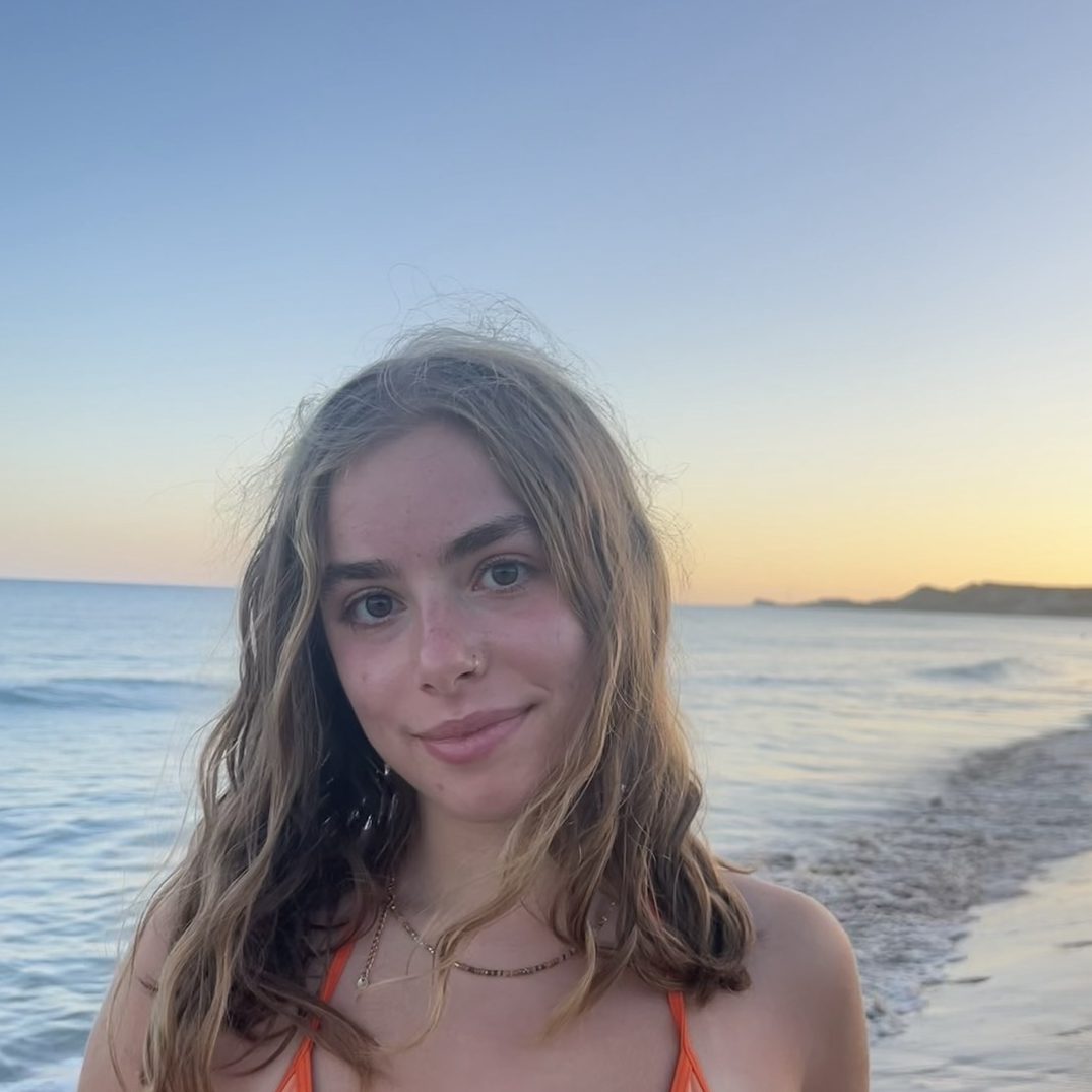 Female student photo wearing a orange top with shoulder length brown hair on a beach.